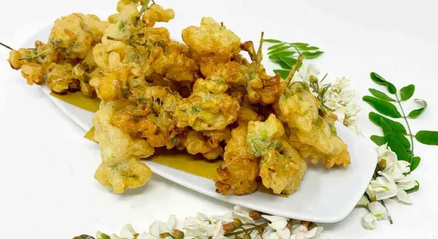 Fiori di acacia fritti, dorati e croccanti, serviti su un piatto bianco con rametti di fiori e foglie fresche.