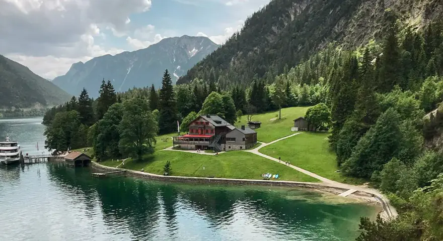 Gaisalm am Achensee: ein Gasthaus mit roten Fensterläden, umgeben von grünen Wiesen und Bergen. Ein Schiff liegt am Steg.