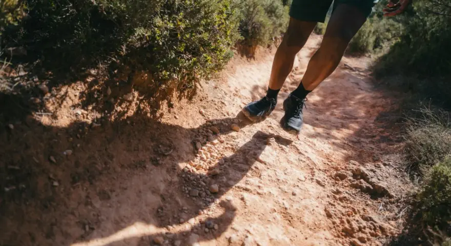 Jambes d'un homme en short et chaussures de trail, s'engageant sur un sentier rocailleux et poussiéreux. L'ombre de son corps s'étire sur le chemin, suggérant un rythme de fast hiking sous le soleil.