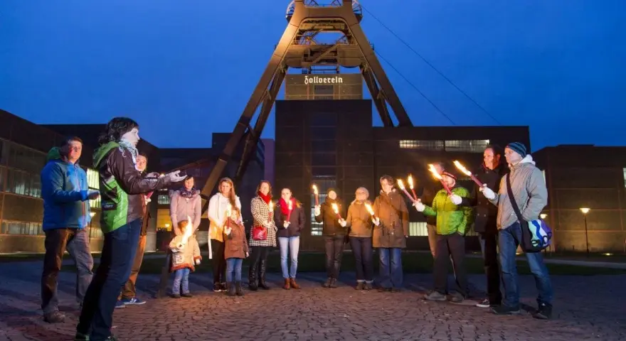 Eine Gruppe von Menschen hält nachts Fackeln vor dem Förderturm der Zeche Zollverein.