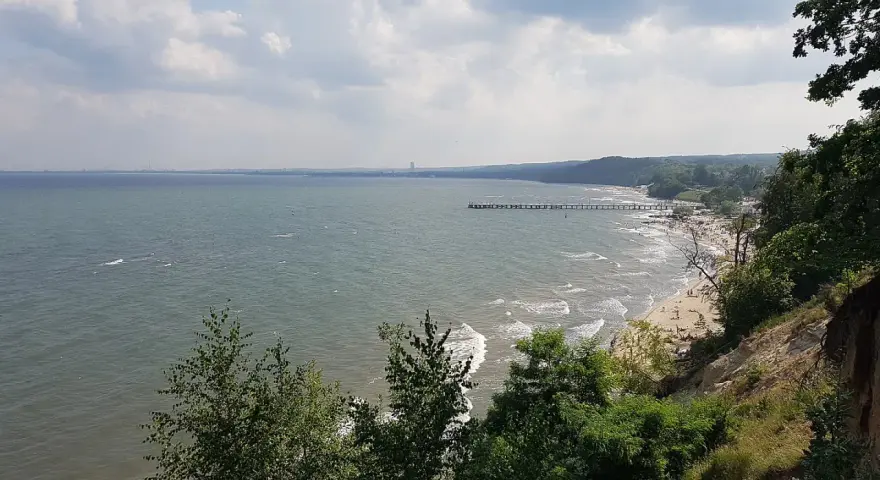 Widok z klifu orłowskiego na morze, plażę z ludźmi i molo. W oddali widać zarys wybrzeża.