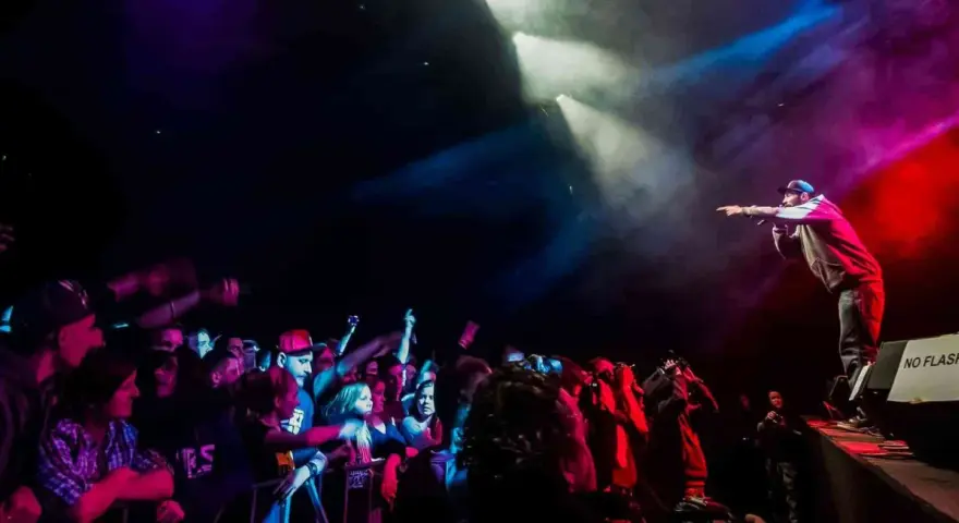 Raper na scenie, w świetle reflektorów, wskazuje palcem na tłum. Koncert w **3 wymiar**ze, pełen energii i emocji.