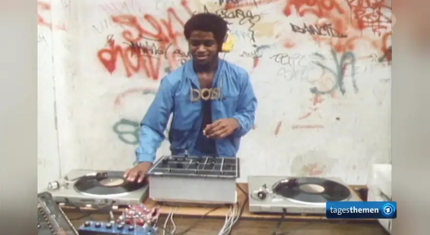 DJ Kool Herc, der oft als derjenige gilt, der Hip Hop erfunden hat, legt auf. Graffiti zieren die Wand im Hintergrund.