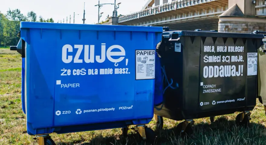Niebieski kontener na makulaturę z napisem "czuję, że coś dla mnie masz. PAPIER" obok czarnego na odpady zmieszane.