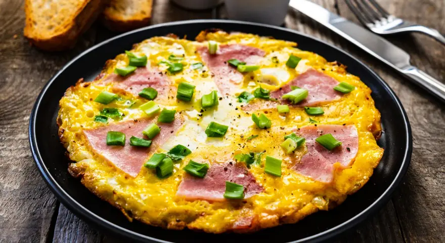 Pyszny omlet z szynką i serem, posypany szczypiorkiem, podany z grzankami. Idealny na śniadanie.