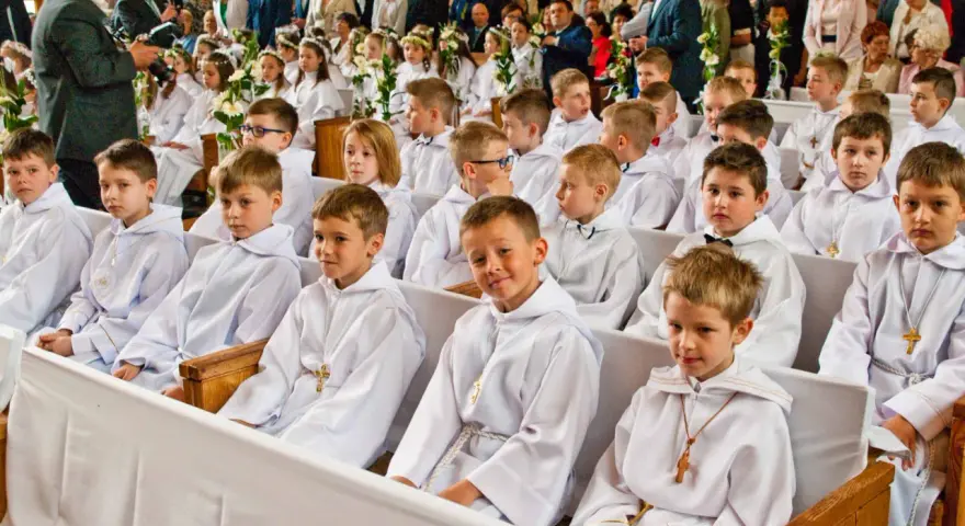 Dzieci w bieli, gotowe na rocznicę komunii świętej, siedzą w kościele. W tle widać zgromadzonych gości i fotografa.