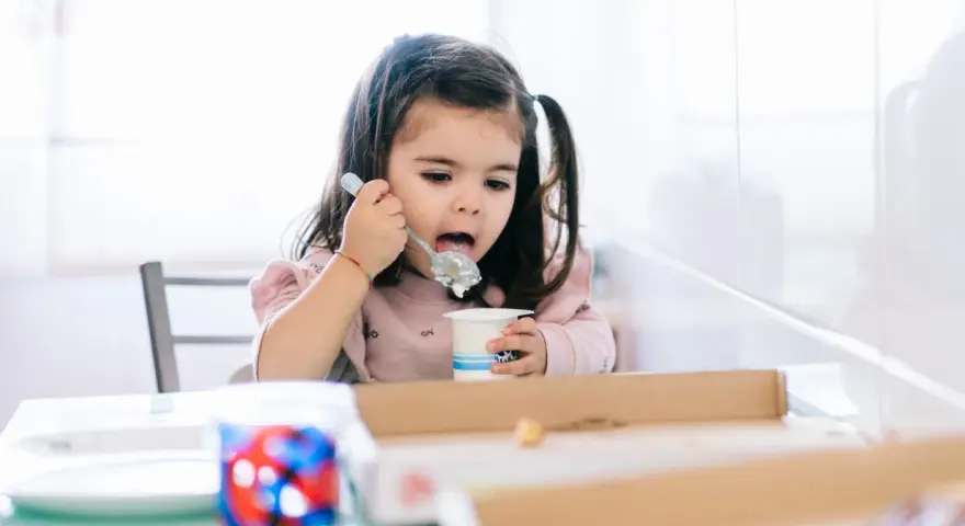 Mała dziewczynka z kitkami je jogurt z kubeczka. Od kiedy dziecko może jeść danonki? Odpowiedź jest prosta: gdy tylko zaczyna jeść pokarmy stałe.