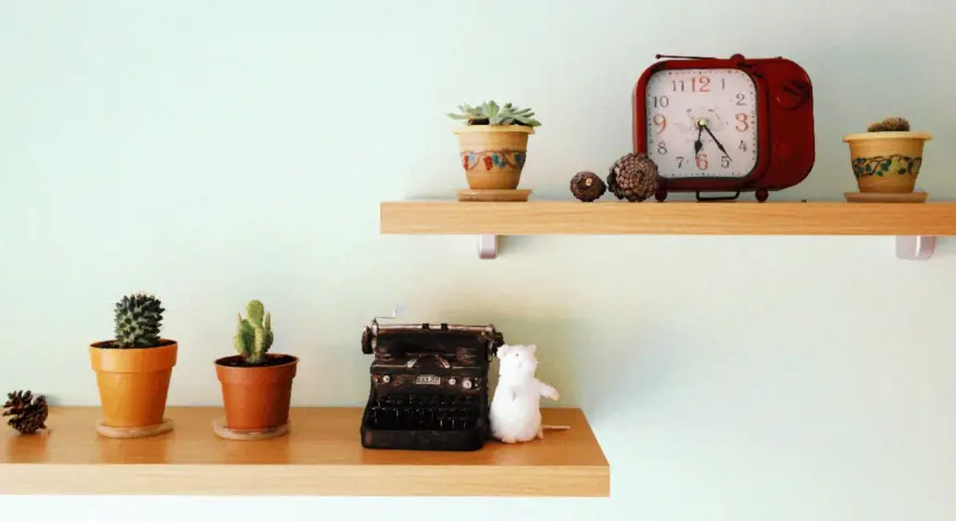 Ideas para decorar baldas salón: dos estantes de madera con cactus, una máquina de escribir antigua, un peluche blanco y un reloj rojo.