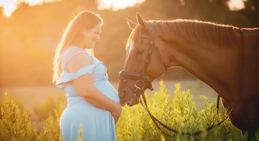 Une femme enceinte en robe bleue caresse un cheval brun au coucher du soleil. L'équitation enceinte est une belle façon de célébrer la maternité.
