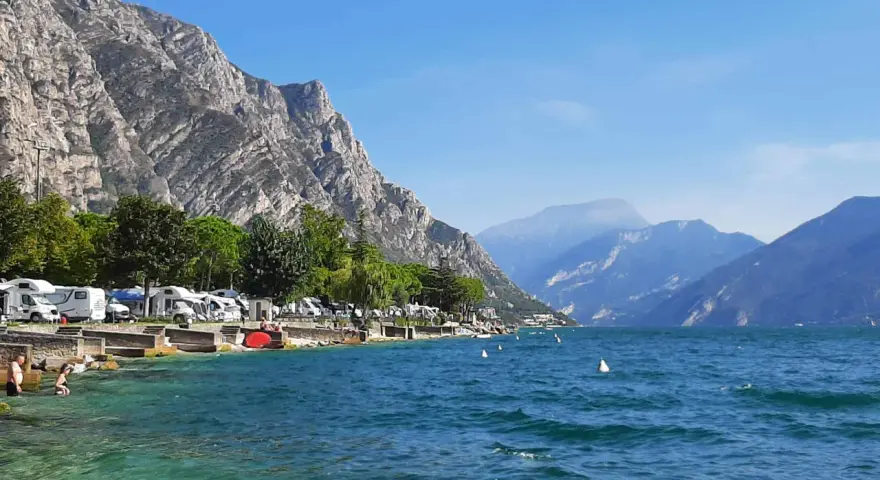 Malowniczy camping nad jeziorem Garda, z kamperami przy brzegu i górami w tle.