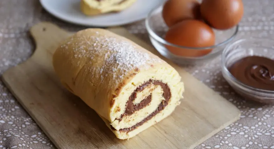 Un delizioso rotolo alla nutella, spolverato di zucchero a velo, pronto per essere gustato. Ingredienti freschi, uova e crema al cioccolato, completano la scena.