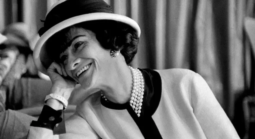 Coco Chanel sorride con un cappello e perle, un'icona di stile che sembra raccontare la storia di quando è nata Coco Chanel.