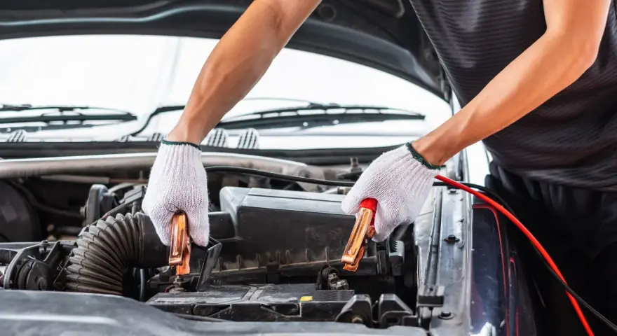 Hands in white gloves connect jumper cables to a car battery. This process helps answer how long does it take to charge a car battery.