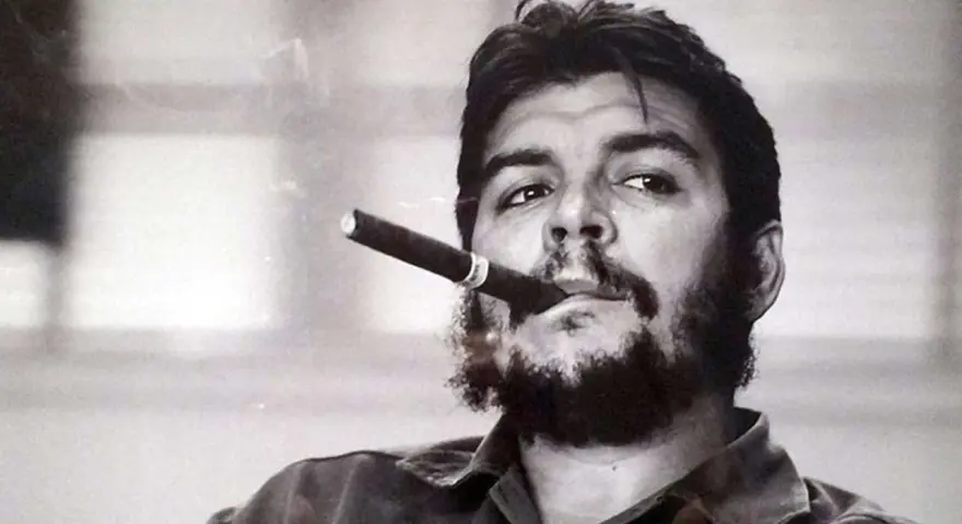 Portrait en noir et blanc de Che Guevara, cigare aux lèvres, regard intense. L'image évoque sa légende, même après sa mort.