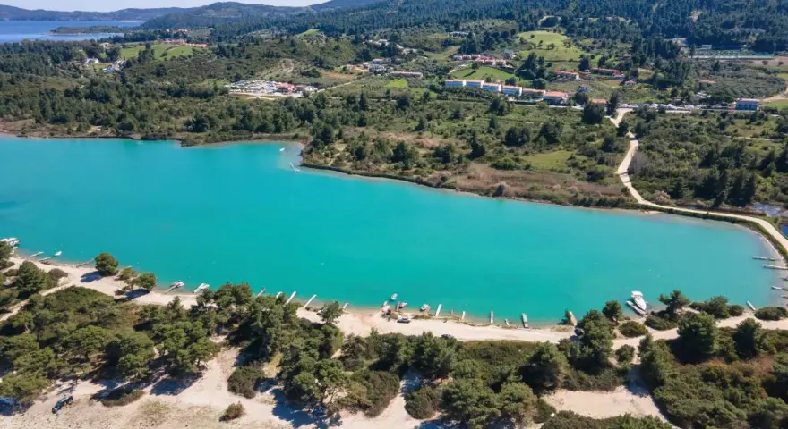 Turkusowa laguna na Kassandrze, otoczona zielonymi wzgórzami i lasem sosnowym. W oddali widać zabudowania i plażę z łodziami.