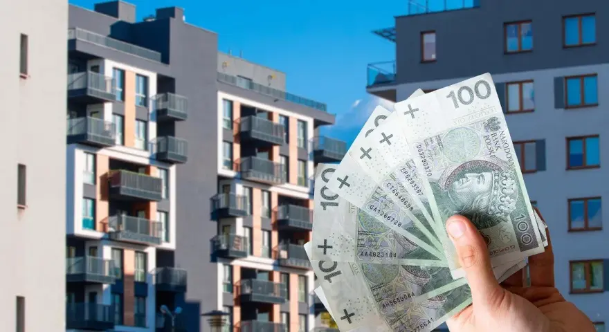 Dłoń trzyma banknoty 100 zł na tle nowoczesnych bloków mieszkalnych. Czy to wyjaśnia, dlaczego ceny najmu w Warszawie i Krakowie przestały rosnąć w pierwszym kwartale 2026?
