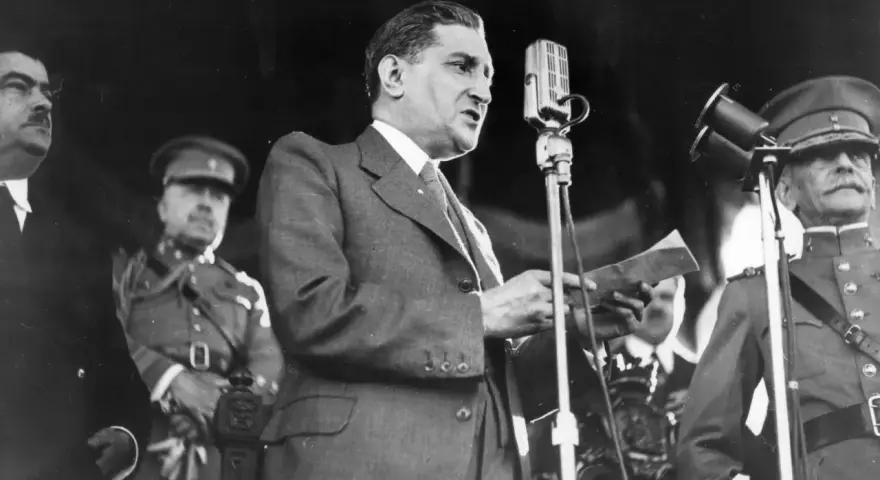 Un homme en costume parle à un microphone, entouré d'officiers en uniforme. Un moment clé de la dictature portugaise de Salazar.