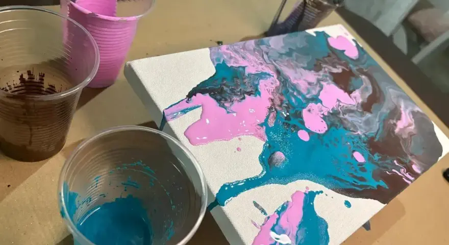 Peinture acrylique sur toile, avec des couleurs vives de rose, bleu sarcelle et marron qui coulent. Des gobelets en plastique contiennent les peintures.