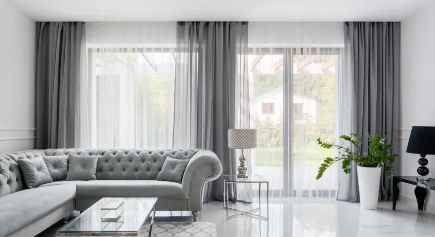 Szary, pikowany narożnik i szklane stoliki tworzą elegancki salon. Duże okna z zasłonami dodają mu **glamuru**.