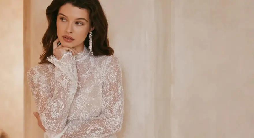 Elegancka, biała sukienka bez dekoltu, zdobiona koronką i cekinami, z długimi rękawami.