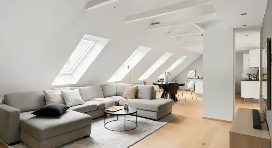 Soggiorno mansardato con divano angolare grigio, tavolino nero e travi a vista moderne dipinte di bianco che creano un'atmosfera luminosa.