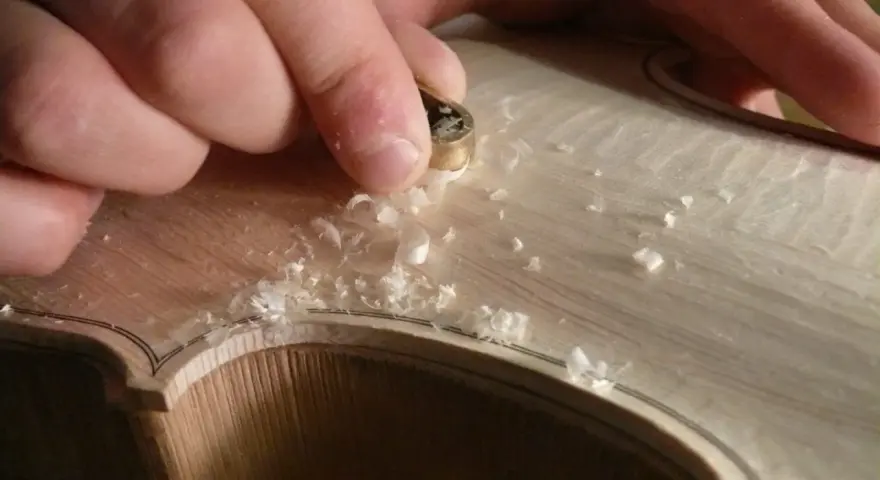Hände bearbeiten mit einem Werkzeug das helle Holz einer Geige, Späne fliegen. Die Frage "aus welchem Holz besteht die Geige" wird hier visuell beantwortet.