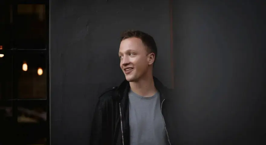 Joris, lächelnd, lehnt an einer dunklen Wand.
