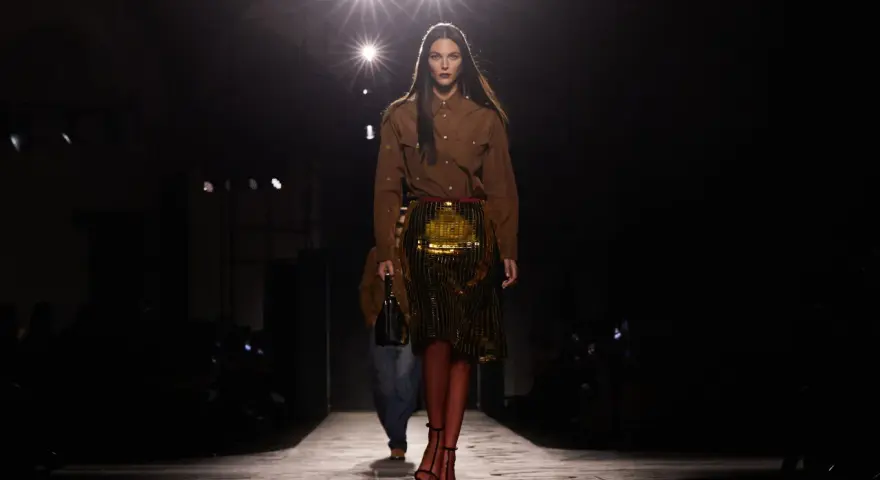 Modella su passerella, con gonna dorata e camicia marrone, un omaggio all'eleganza senza tempo di Prada, fondatore di un impero della moda.
