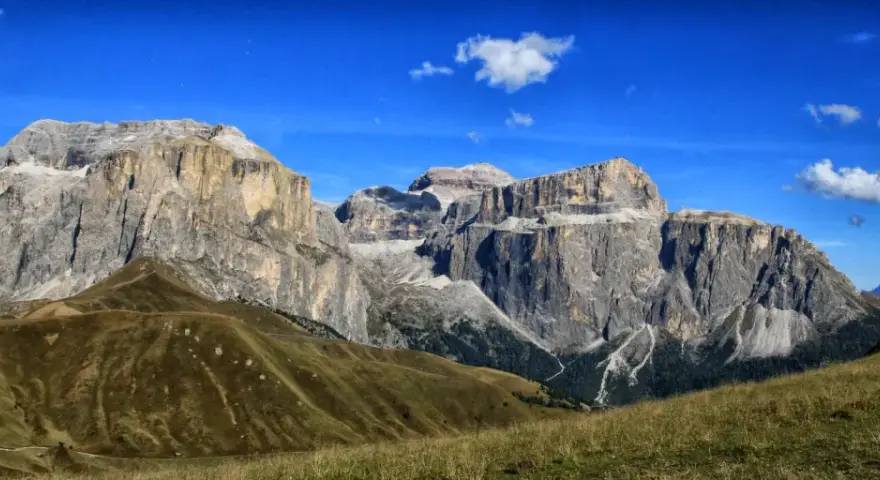 Vaste cime rocciose del gruppo Sella Dolomiti sotto un cielo azzurro con nuvole bianche. Prati verdi e sentieri serpeggiano sui pendii.