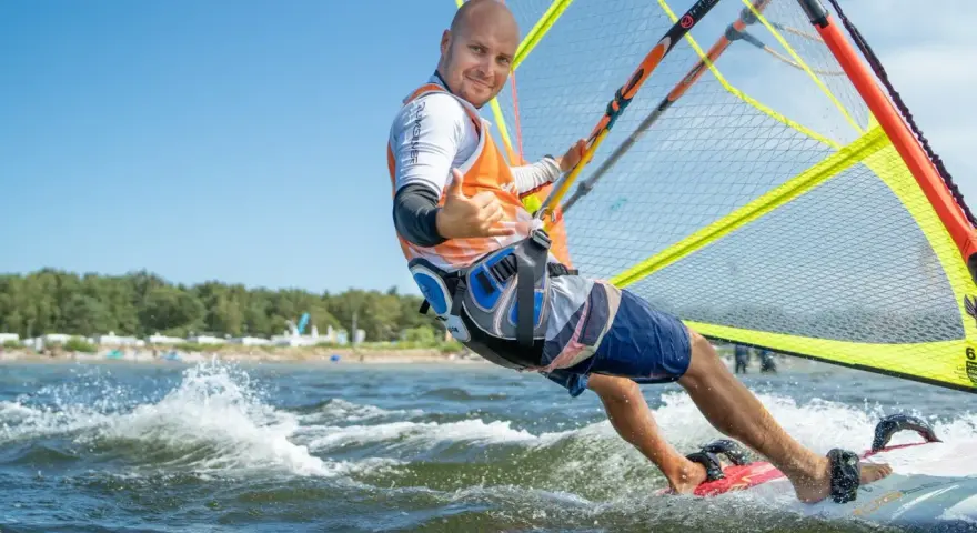 Mężczyzna na desce windsurfingowej, uśmiecha się i pokazuje kciuk w górę.