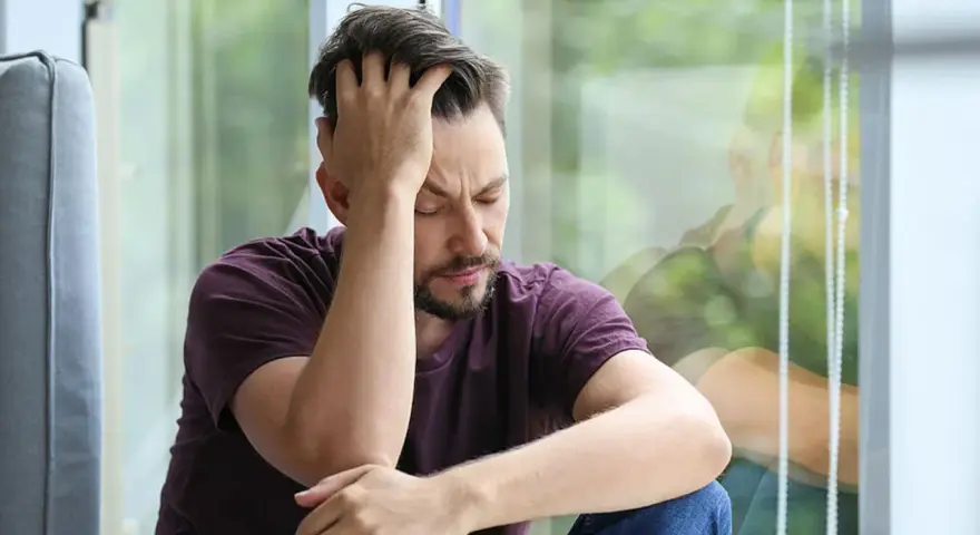 Mann sitzt mit geschlossenen Augen und der Hand am Kopf vor einem Fenster.