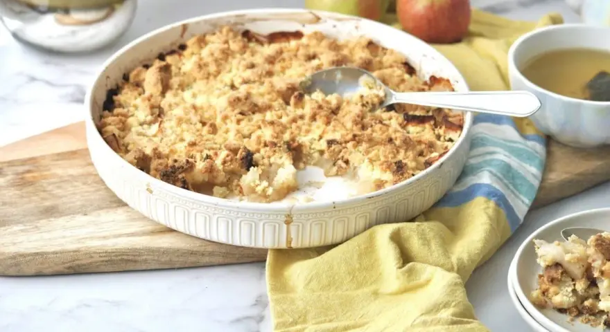 Ein goldbrauner Apfel-Crumble in einer Auflaufform, bereit zum Servieren. Daneben eine Tasse Tee und frische Äpfel.