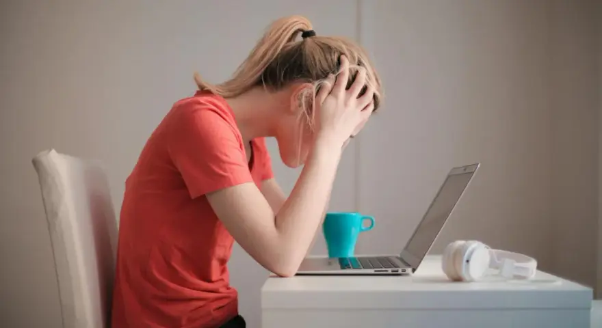 Kobieta z rękami na głowie, przy laptopie, z kubkiem i słuchawkami. Cierpi na uzależnienie od stresu, czuje się przytłoczona.