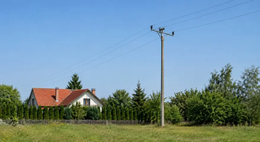 Słup na działce, obok domu z czerwoną dachówką i żywopłotem. W tle zielone drzewa i błękitne niebo.