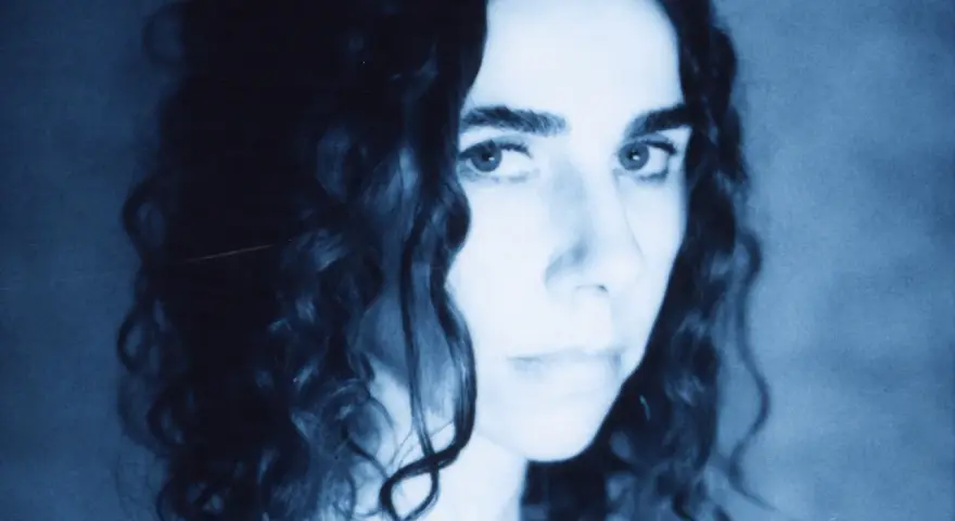 PJ Harvey, z kręconymi włosami, patrzy intensywnie. Jej twarz, w odcieniach niebieskiego, zdaje się opowiadać historię z "I Inside the Old I Dying".