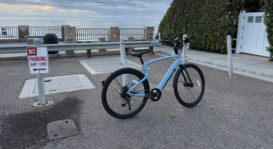 A light blue electric city bike is parked on asphalt near a "No Parking" sign, with the ocean and cloudy sky in the background.