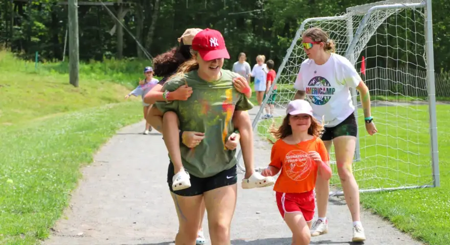 Mädchen haben Spaß bei einem Sommercamp in den USA. Eine trägt die andere auf dem Rücken, während sie lachend über einen Weg laufen.