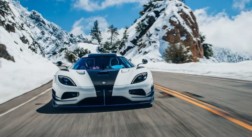 Biały Koenigsegg Agera RS z czarnym pasem na masce pędzi po ośnieżonej górskiej drodze.