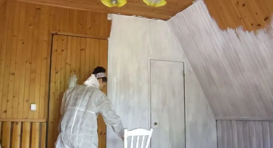 Osoba w kombinezonie maluje lakierowaną boazerię bez szlifowania na biało. Widać postęp prac na ścianie i suficie.
