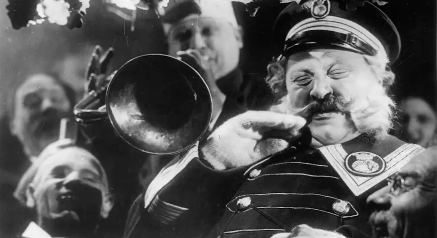 Un uomo con baffi e barba folta suona una tromba, circondato da volti sorridenti. Un'atmosfera da kammerspiel, un dramma intimo e intenso.