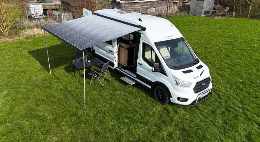 Vit husbil, Karmann Dexter, med utdraget markis och campingmöbler på gräs.