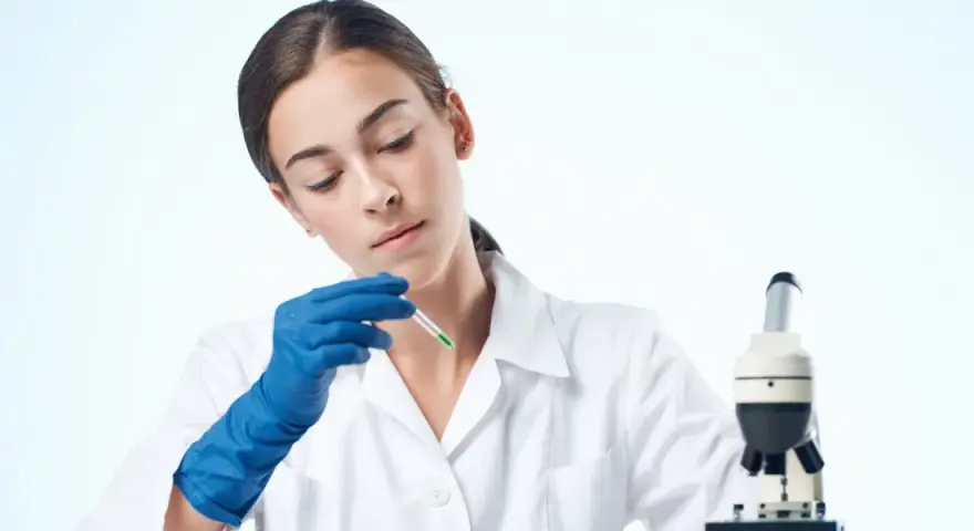 Giovane tecnologo alimentare in camice bianco, con guanti blu, analizza un campione con un contagocce accanto a un microscopio.