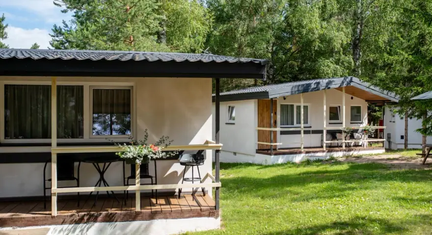 Dwa białe domki kempingowe w lesie, z werandami i meblami ogrodowymi. Idealne miejsce dla każdego **camping wagabunda**.