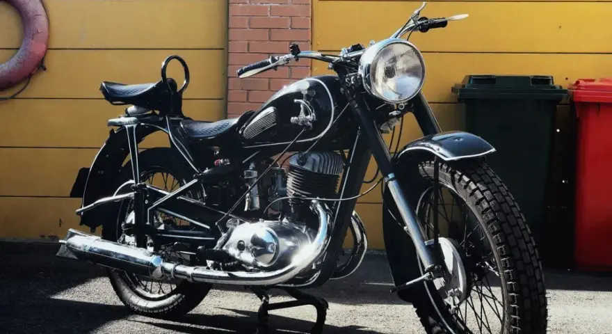 Klasyczny czarny motocykl, model iż 49, lśni w słońcu. Obok żółta brama i ceglana ściana.