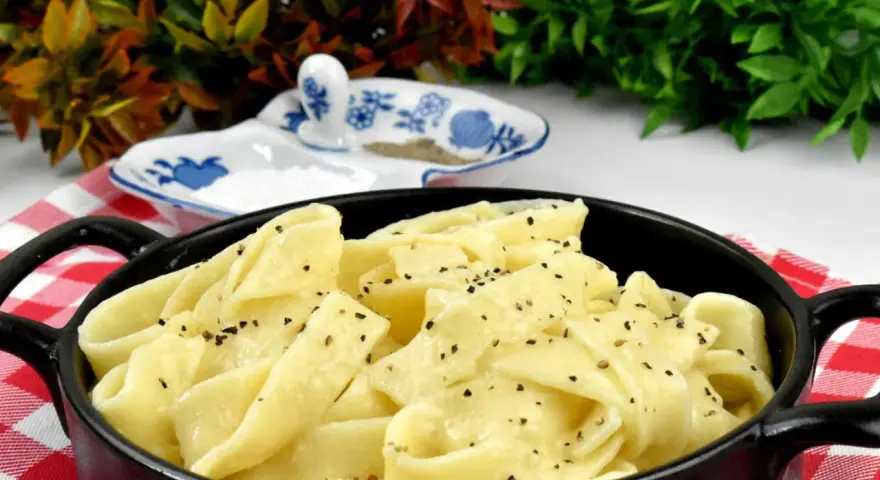 Pappardelle w kremowym sosie alfredo, posypane świeżo mielonym pieprzem, podane w czarnym naczyniu na czerwono-białej kratce.