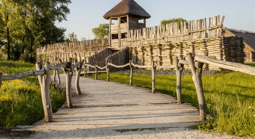 Drewniana kładka prowadzi do grodziska, jednego z historycznych miejsc w Polsce. Widać palisadę i wieżę obronną.