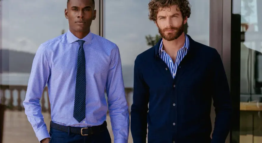 Dos hombres posan. Uno lleva una camisa de vestir azul con corbata, la camisa por dentro. El otro, una camisa de rayas azules y blancas con cárdigan azul oscuro, la camisa por fuera.