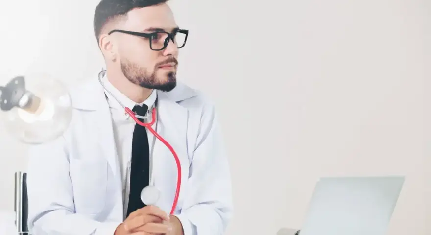Lekarz medycyny pracy w okularach, z czerwonym stetoskopem, siedzi przy biurku z laptopem. Jakie badania wykonuje?