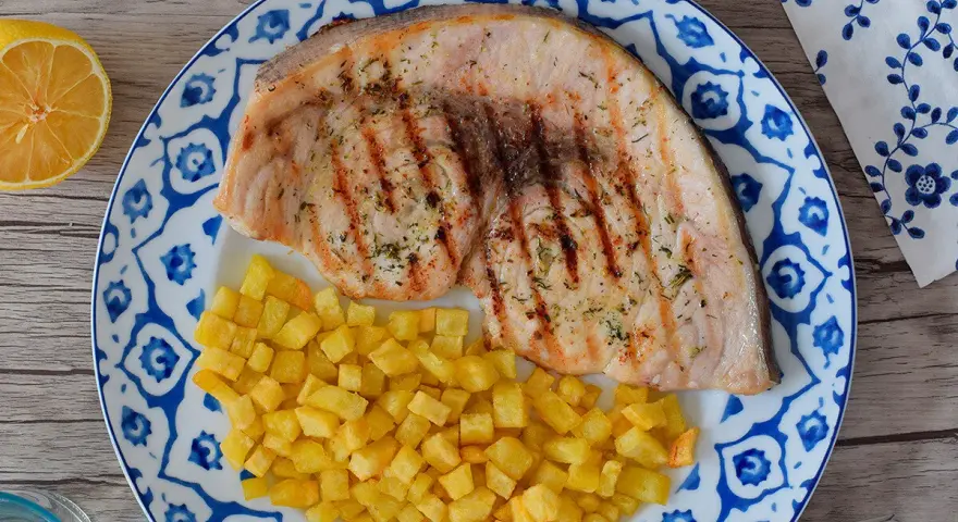 Delicioso emperador a la plancha con patatas fritas crujientes, servido en un plato con diseño azul y blanco. Un toque de limón completa esta apetitosa presentación.