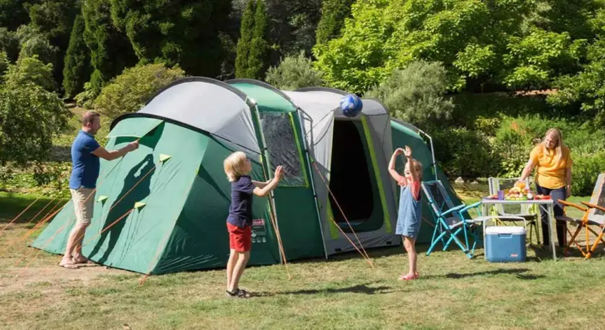 Radosny camping rodzinny: tata rozkłada namiot, dzieci bawią się piłką, mama przygotowuje posiłek.